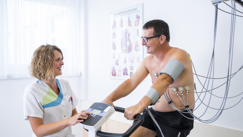 herbst-00194202208254711, © Robert Herbst Ein Mann auf einem Ergometer mit Elektroden am Körper, betreut von einer lächelnden Frau in medizinischer Kleidung.