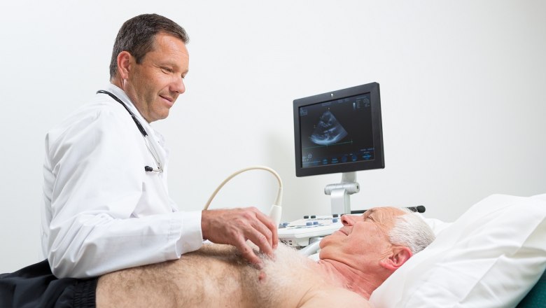 Echokardiographie, © Werner Harrer Ein Arzt führt eine Echokardiographie bei einem älteren Mann durch, der auf einer Liege liegt. Ein Monitor zeigt das Ultraschallbild des Herzens.