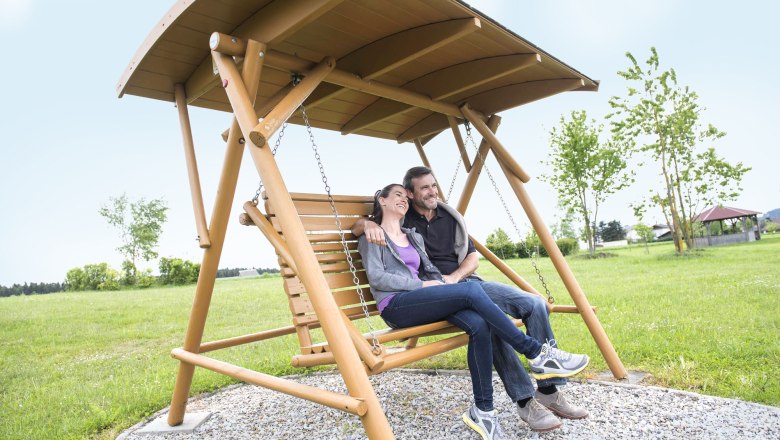 Wohlfühlen in der Entspannungsschaukel, © Xundheitswelt Ein Paar sitzt entspannt auf einer Holzschaukel im Freien.
