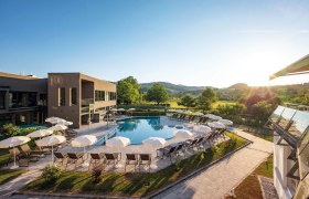Außenansicht, © Hannes Dabernig Außenanlage eines Hotels mit Pool, Sonnenschirmen, Liegestühlen, umgeben von grünen Wiesen und Bäumen.