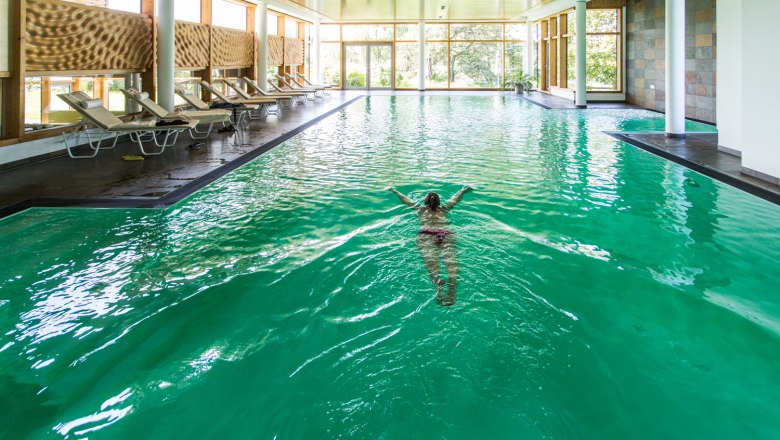 Hallenbad, © Gesundheitshotel Klosterberg Person schwimmt in einem Hallenbad mit grünen Wasser und Liegestühlen am Rand.