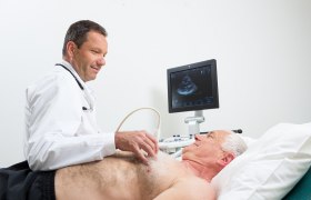 Ein Arzt führt eine Echokardiographie bei einem älteren Mann durch, der auf einer Liege liegt. Ein Monitor zeigt das Ultraschallbild des Herzens.