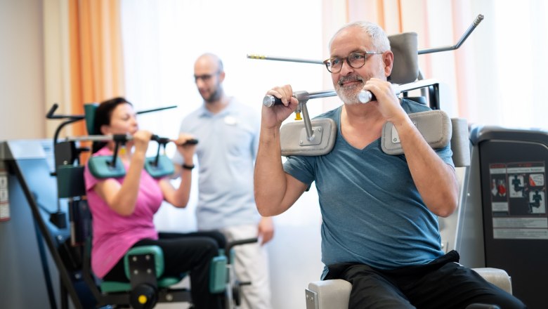Zwei Personen trainieren an Fitnessger&auml;ten, w&auml;hrend ein Trainer zuschaut.