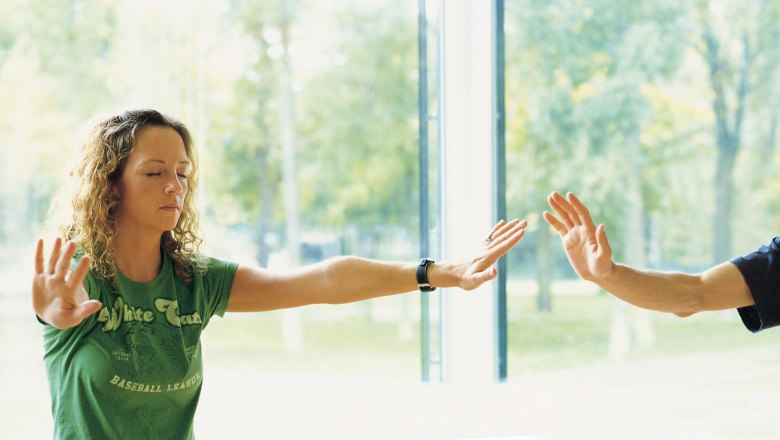 Zwei Personen mit ausgestreckten Armen in einem hellen Raum, eine Frau mit geschlossenen Augen.