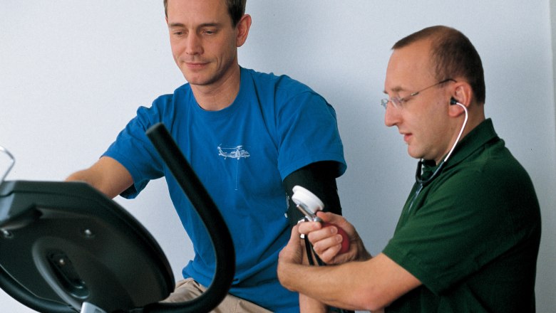 Ein Mann auf einem Ergometer wird von einem Arzt mit einem Blutdruckmessgerät untersucht.