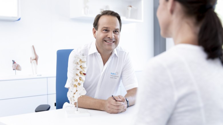 Ein Arzt im weißen Kittel spricht mit einer Patientin in einem modernen Behandlungsraum.