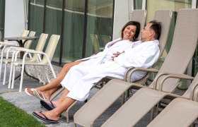 Ein Paar in weißen Bademänteln entspannt auf Liegestühlen im Freien.