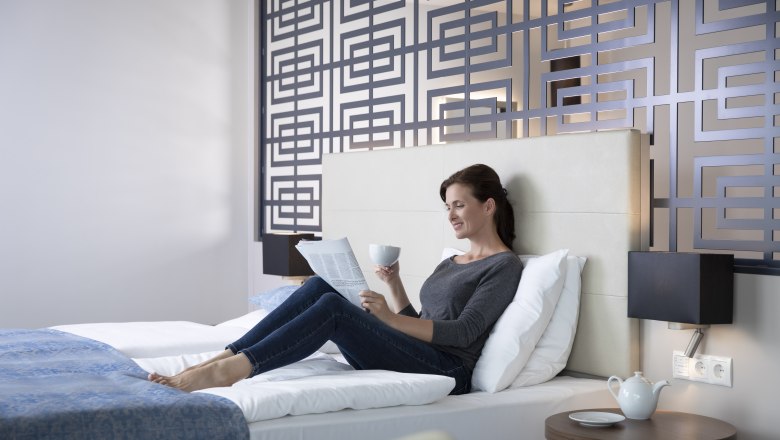 Frau entspannt auf einem Bett in einem modernen Hotelzimmer, liest eine Zeitung und hält eine Tasse. Dekorative Wand im Hintergrund.