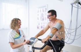 Ein Mann auf einem Ergometer mit Elektroden am Körper, betreut von einer lächelnden Frau in medizinischer Kleidung.