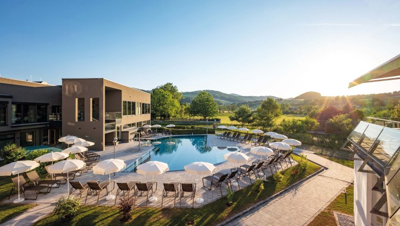 Außenansicht, © Hannes Dabernig Außenanlage eines Hotels mit Pool, Sonnenschirmen, Liegestühlen, umgeben von grünen Wiesen und Bäumen.